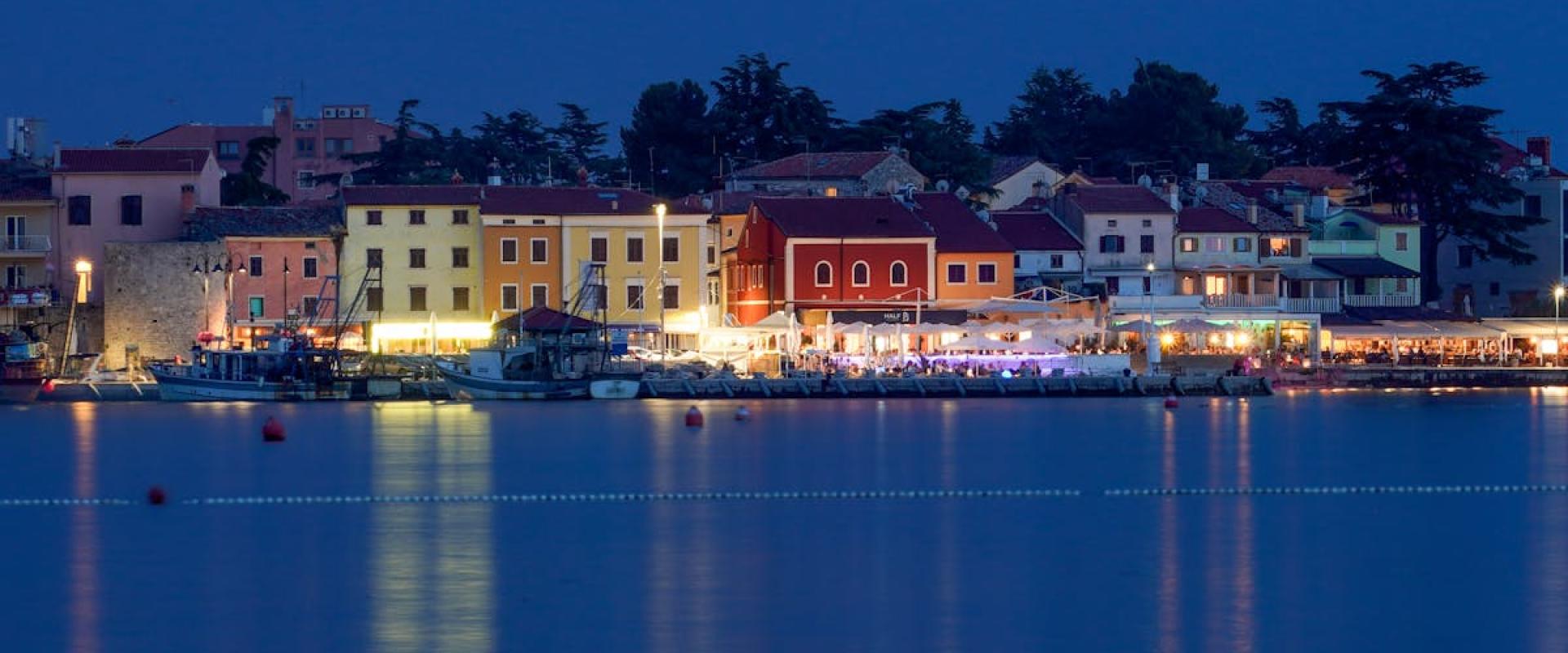 Illuminated harbor with colorful houses in Novigrad, Croatia at night, reflecting in calm water.