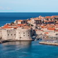 Aerial view of the historic walled city of Dubrovnik, Croatia, showing its distinctive red-tiled roofs, stone fortifications, the Old Port with boats, and the surrounding Adriatic Sea under a cloudy sky.