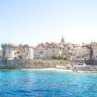A stone-walled medieval town with towers and red-tiled roofs situated on a small island or peninsula, viewed from the sea under a clear blue sky.