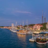 A harbor scene at dusk showing several traditional wooden boats and yachts docked along a waterfront promenade with historic buildings, including a prominent stone tower and red-roofed structures, under a twilight sky.