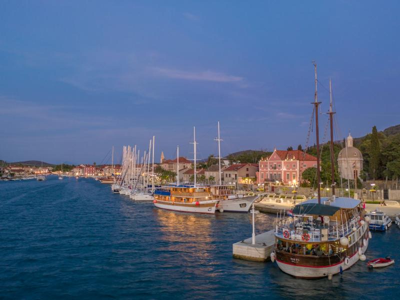 A harbor scene at dusk showing several traditional wooden boats and yachts docked along a waterfront promenade with historic buildings, including a prominent stone tower and red-roofed structures, under a twilight sky.