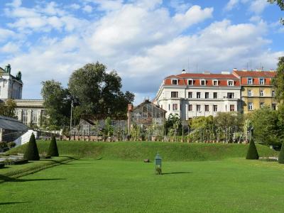 The image shows a large grassy park with manicured lawns, conical topiary trees, and several glasshouse structures in the background, with a prominent white building featuring a red-tiled roof on the right side.