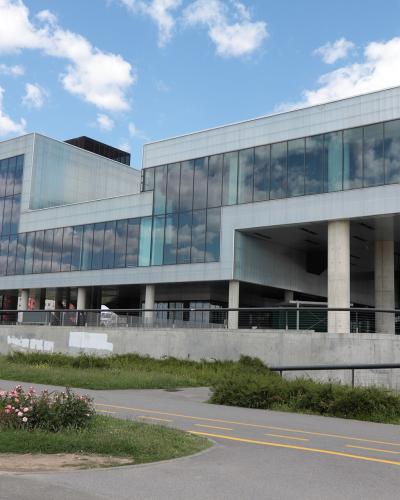A modern, angular building with extensive glass facades and concrete supports, featuring a cyclist on a paved path in the foreground under a partly cloudy sky.