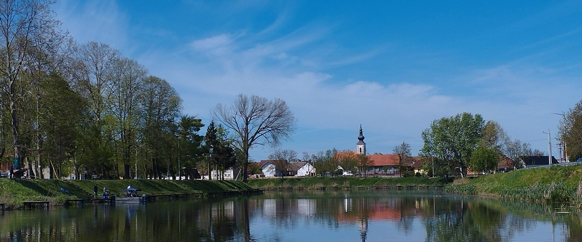 A calm river or canal reflecting a blue sky, with trees lining the banks and a village featuring a church steeple in the background.