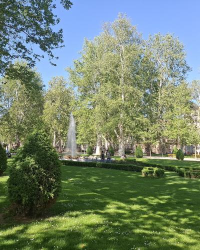 A sunlit public park with manicured lawns, tall poplar trees, sculpted shrubs, and active water fountains, with people walking and sitting on benches under a clear blue sky.