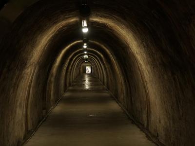 A long, arched concrete tunnel with a central row of overhead lights and a wet-looking floor leading to a bright exit at the far end.
