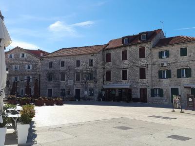 A wide, paved stone square in a Mediterranean town, framed by historic limestone buildings with shuttered windows and red-tiled roofs, featuring closed cafe umbrellas and outdoor seating in the foreground.