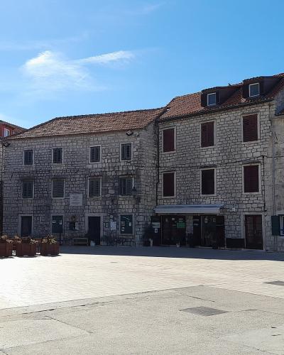 A wide, paved stone square in a Mediterranean town, framed by historic limestone buildings with shuttered windows and red-tiled roofs, featuring closed cafe umbrellas and outdoor seating in the foreground.
