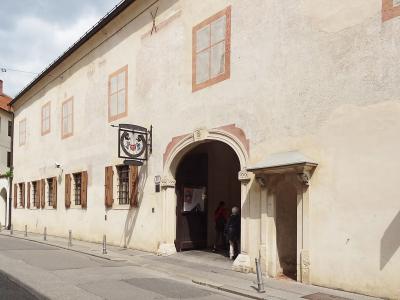 The image shows the exterior of a historic building with a large arched entrance, a hanging sign featuring a lyre and the letters "MGZ", and a street number "20" visible on the wall.