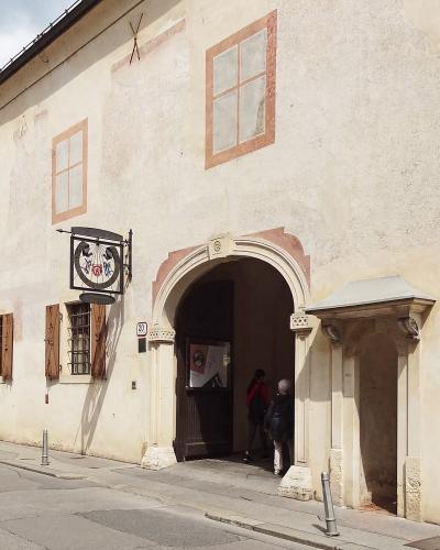 The image shows the exterior of a historic building with a large arched entrance, a hanging sign featuring a lyre and the letters "MGZ", and a street number "20" visible on the wall.