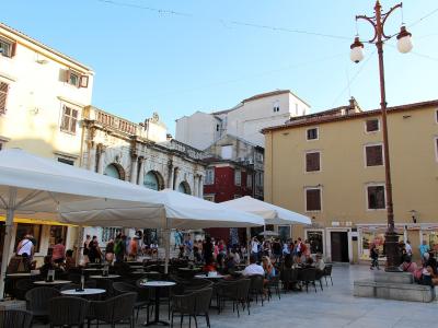 An outdoor plaza with cafe seating under white umbrellas, surrounded by historic buildings including a prominent Roman triumphal arch (the Arch of the Sergii) and a distinctive ornate lamppost.