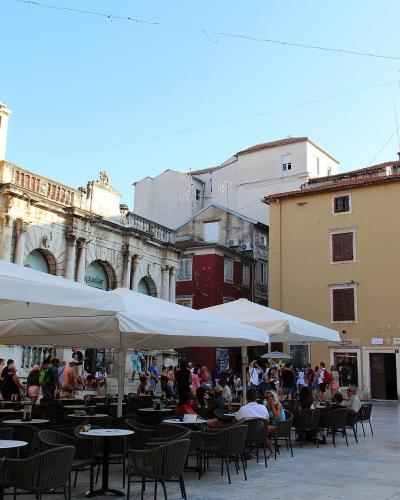 An outdoor plaza with cafe seating under white umbrellas, surrounded by historic buildings including a prominent Roman triumphal arch (the Arch of the Sergii) and a distinctive ornate lamppost.
