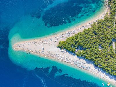 An aerial photograph of a distinctive V-shaped sandy peninsula beach crowded with people and umbrellas, bordered by a dense pine forest on one side and clear blue-green sea on the other.