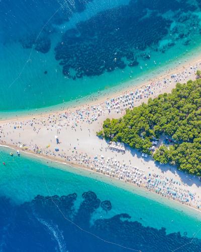 An aerial photograph of a distinctive V-shaped sandy peninsula beach crowded with people and umbrellas, bordered by a dense pine forest on one side and clear blue-green sea on the other.
