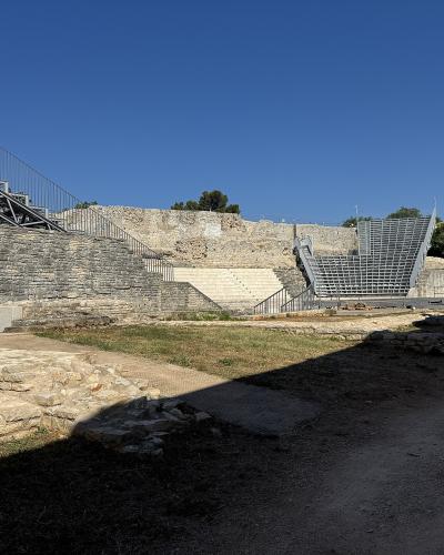 An ancient stone theatre ruin featuring modern metal staircases and seating structures installed within the archaeological remains under a clear blue sky.