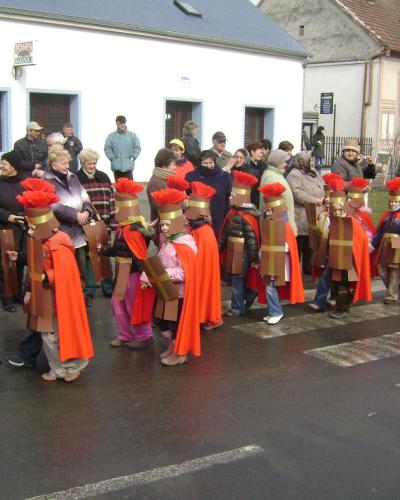 A group of children and adults in homemade Roman soldier costumes with red capes and cardboard armor marching in a street parade while spectators watch from the sidewalk.