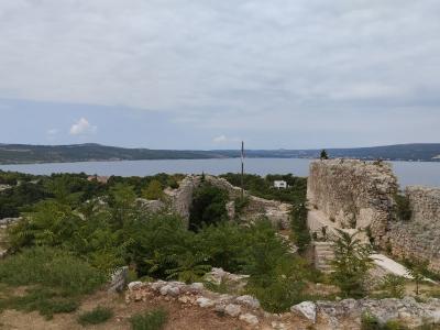 A view of ancient stone ruins and fortification walls on a hillside overlooking a body of water, with a Croatian flag flying on a pole in the middle distance.