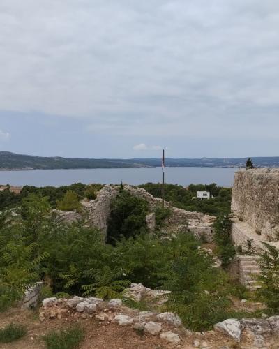 A view of ancient stone ruins and fortification walls on a hillside overlooking a body of water, with a Croatian flag flying on a pole in the middle distance.