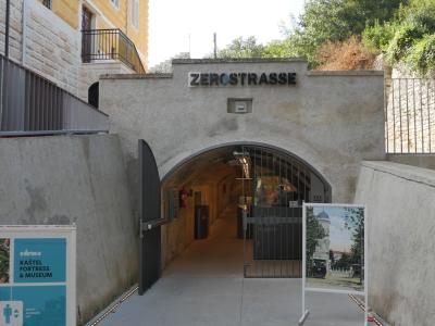 A concrete arched entrance to an underground tunnel with the name "ZEROSTRASSE" mounted above the archway, flanked by informational signage including a blue sign for "KAŠTEL FORTRESS & MUSEUM" and a historical photo display.