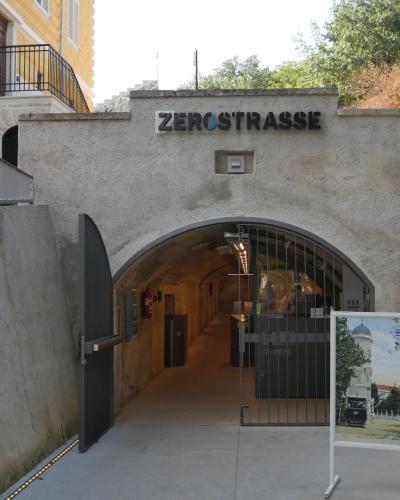 A concrete arched entrance to an underground tunnel with the name "ZEROSTRASSE" mounted above the archway, flanked by informational signage including a blue sign for "KAŠTEL FORTRESS & MUSEUM" and a historical photo display.