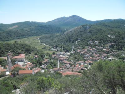 A high-angle landscape view of a coastal village with terracotta-roofed buildings nestled in a green valley, surrounded by forested hills and mountains under a clear sky.