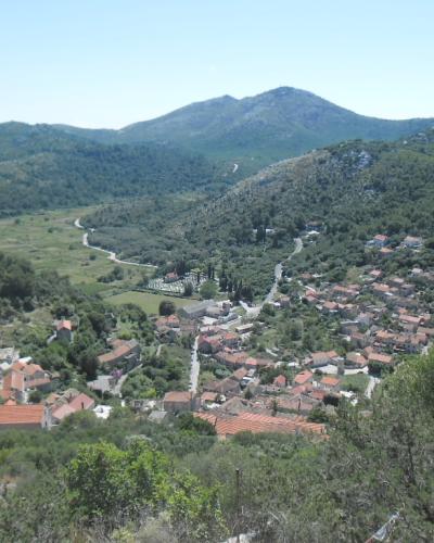A high-angle landscape view of a coastal village with terracotta-roofed buildings nestled in a green valley, surrounded by forested hills and mountains under a clear sky.