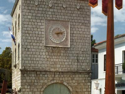 A stone clock tower building with a large sundial on its facade, situated in a paved town square next to a fountain and a flagpole flying a banner.