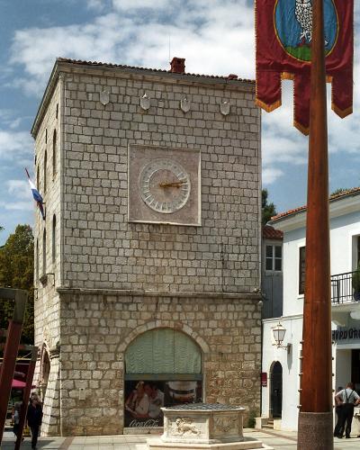 A stone clock tower building with a large sundial on its facade, situated in a paved town square next to a fountain and a flagpole flying a banner.