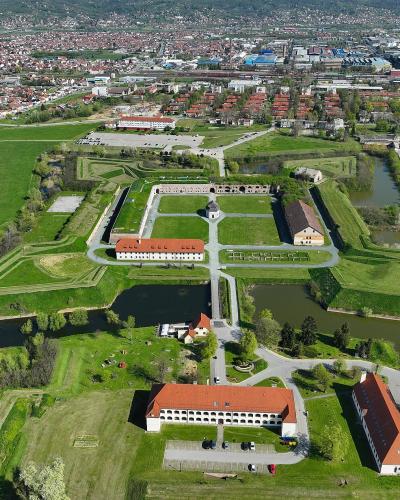 An aerial view of a large, star-shaped historical fortress with surrounding moats, grassy ramparts, and several long buildings with red-tiled roofs, situated within a modern city.