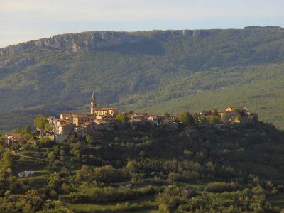 A wide landscape shot of a medieval hilltop town with terracotta-roofed buildings and a prominent church tower, set against a backdrop of forested hills and a distant rocky plateau.