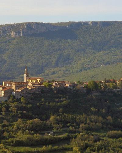 A wide landscape shot of a medieval hilltop town with terracotta-roofed buildings and a prominent church tower, set against a backdrop of forested hills and a distant rocky plateau.