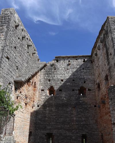 A low-angle view of a medieval stone fortress with two prominent towers and crenellated walls, featuring small arched windows and exposed brickwork, set against a blue sky with some greenery in the foreground.