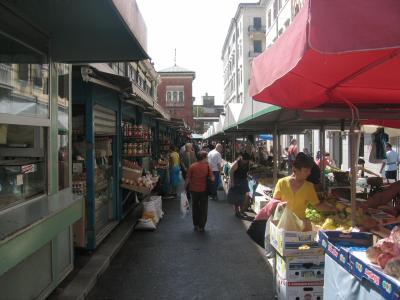 An outdoor market scene with stalls on both sides of a paved walkway, featuring a red umbrella on the right, a brick building with arched windows in the background, and shoppers browsing produce and goods.