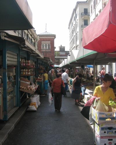 An outdoor market scene with stalls on both sides of a paved walkway, featuring a red umbrella on the right, a brick building with arched windows in the background, and shoppers browsing produce and goods.