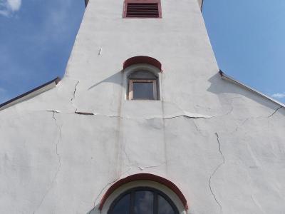 A low-angle shot of a white church facade with a tall tower, featuring a red-tiled spire, a louvered vent, and arched windows, with visible cracks in the plaster.