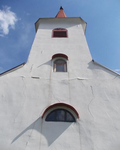 A low-angle shot of a white church facade with a tall tower, featuring a red-tiled spire, a louvered vent, and arched windows, with visible cracks in the plaster.