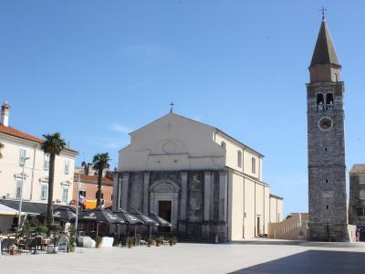The image shows a large, paved public square featuring a prominent church with a stone facade and a tall, separate stone bell tower (campanile) to the right, set against a clear blue sky.