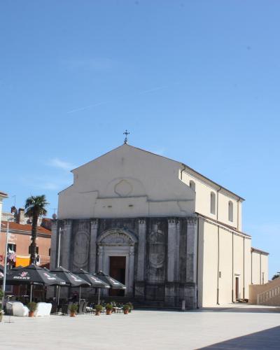 The image shows a large, paved public square featuring a prominent church with a stone facade and a tall, separate stone bell tower (campanile) to the right, set against a clear blue sky.