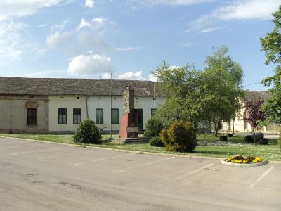 A paved parking lot in front of a long, single-story building with a tiled roof, featuring a central stone and red monument, several flagpoles, and landscaped greenery under a blue sky.