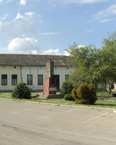 A paved parking lot in front of a long, single-story building with a tiled roof, featuring a central stone and red monument, several flagpoles, and landscaped greenery under a blue sky.