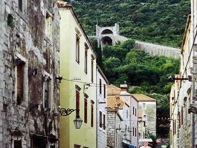 A narrow, cobblestone street (Široka ulica) in Ston, Croatia, looking uphill towards the historic Ston Walls with a distinctive arched gateway, flanked by old stone and plastered buildings, with a white car parked on the left and outdoor restauran