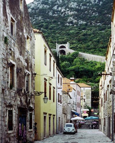 A narrow, cobblestone street (Široka ulica) in Ston, Croatia, looking uphill towards the historic Ston Walls with a distinctive arched gateway, flanked by old stone and plastered buildings, with a white car parked on the left and outdoor restauran