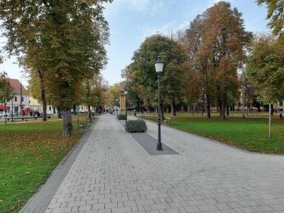 A paved pedestrian walkway lined with trees showing autumn foliage, benches, and vintage-style lampposts in a town square or park setting.