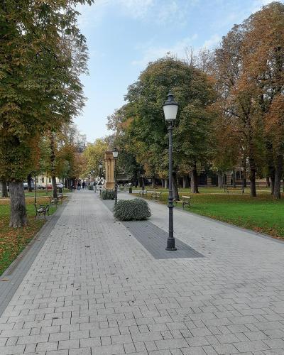 A paved pedestrian walkway lined with trees showing autumn foliage, benches, and vintage-style lampposts in a town square or park setting.