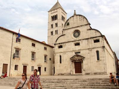 A large, light-colored stone church with a tall bell tower and a distinctive facade featuring a rose window and a pedimented entrance, situated next to an adjacent building flying a Croatian flag.