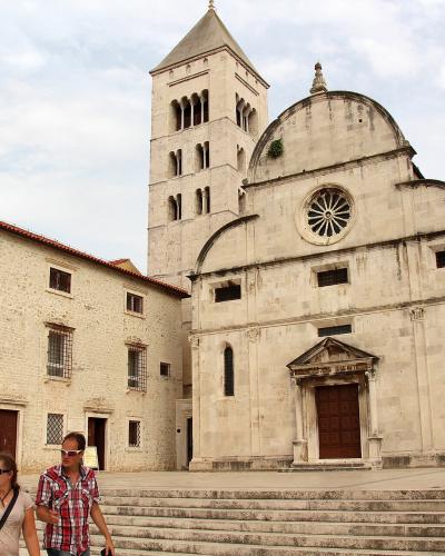 A large, light-colored stone church with a tall bell tower and a distinctive facade featuring a rose window and a pedimented entrance, situated next to an adjacent building flying a Croatian flag.