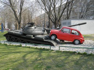 A military tank with the number 20442 on its turret is mounted on a pedestal next to a red Fiat 500 car, set in a grassy park area with trees and buildings in the background.