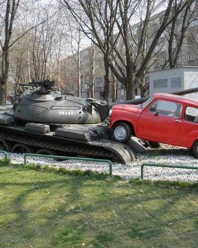 A military tank with the number 20442 on its turret is mounted on a pedestal next to a red Fiat 500 car, set in a grassy park area with trees and buildings in the background.