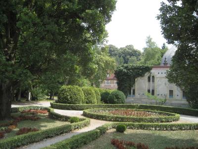 A manicured garden with low hedges and flowerbeds in the foreground, with a historic building featuring a dome and Moorish-style architectural details visible in the background amidst trees.