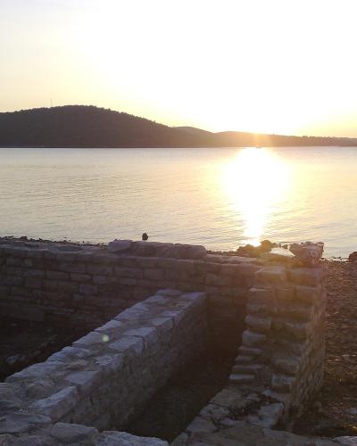 Low stone ruins of an ancient structure situated on a pebble beach next to the sea during sunset.